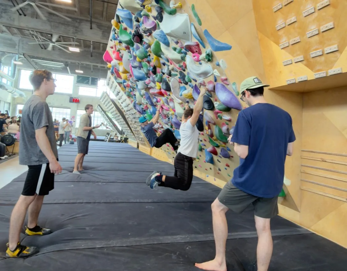 A Vertical Scramble event: two climbers hanging on an overhanging bouldering wall while a reader and other players watch from the mats